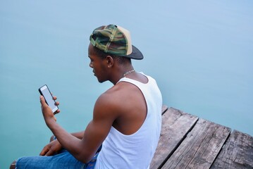 Afro Caribbean Man Sitting By The Dock  On A Rustic Deck With His Mobile Phone.