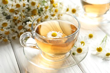 Aromatic tea in glass cup and chamomile flowers on white wooden table, closeup