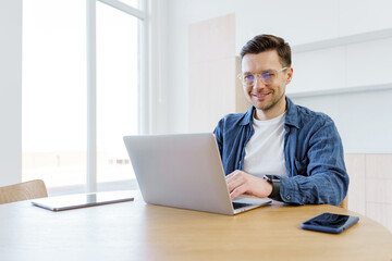Fototapeta premium Smiling young man working on laptop in a cozy, modern workspace while sipping coffee on a bright morning