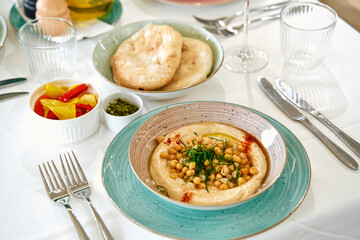 Delicious Spread of Hummus with Chickpeas, Fresh Herbs, and Bread Surrounded by Colorful Pickles and Olive Oil on a Tastefully Set Dining Table