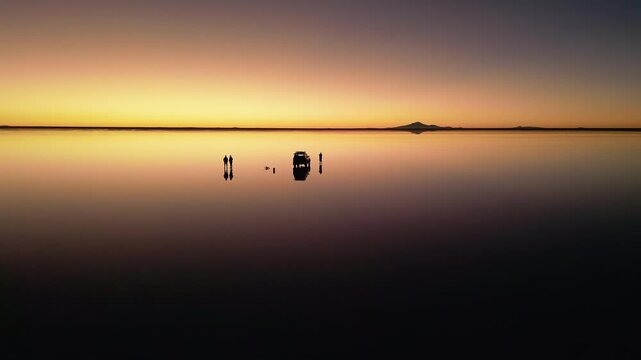 Drone footage circling 4x4 vehicle and two people on chairs at salt flats during sunset. Perfect mirror reflection on water surface. Mountains silhouetted on horizon at dusk.