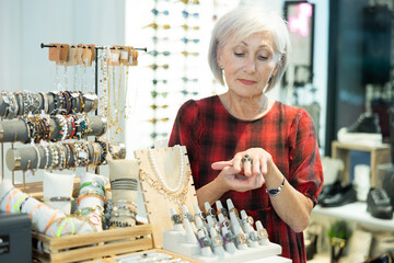 Cheerful elderly female fashionista choosing and trying on various rings while standing near...