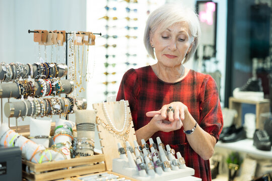 Cheerful elderly female fashionista choosing and trying on various rings while standing near display with costume jewelry in boutique ..