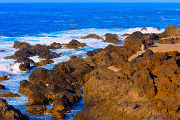Dramatic coastal scene with powerful waves against volcanic rock formations. Coastal view of a rocky beach with turquoise water and a vibrant blue sky