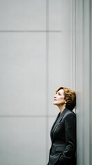 Thoughtful woman in dark gray suit, leaning against a light gray wall, looking upward. Natural light illuminates her.