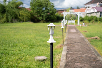 White garden lamps line a brick pathway in a sunny open field, leading to a white arch and bench. A...