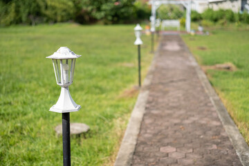 White garden lamps line a brick pathway in a sunny open field, leading to a white arch and bench. A...