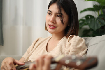Side view, Beautiful Asian woman enjoys playing music with ukulele while sitting on the sofa in...