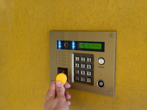 Closeup of a person's hand holding a yellow rfid key fob near reader access with camera, keypad, screen showing "please come in" sign on yellow wall, security, access control, contactless entry system