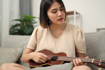Portrait of Beautiful Asian woman enjoys playing music with ukulele while sitting on the sofa in living room at home. Enjoying Weekend.