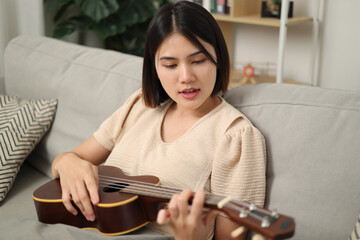 Portrait of Beautiful Asian woman enjoys playing music with ukulele while sitting on the sofa in living room at home. Enjoying Weekend.