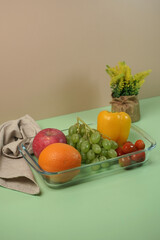 Fresh fruits and vegetables in a glass dish. Healthy food concept with apple, orange, grapes, bell pepper, and tomatoes on a green background.