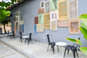 Blurred of empty white tables and black chairs set in garden. Outdoor seating for restaurant, Cafe or coffee shop. Plants, green leaves And wall. Background. Old windows hanging on grey or gray wall. 