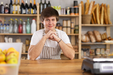 In sales area of store, guy seller is waiting for visitors. Employee in apron uniform work in grocery store, meeting visitors and serves buyers. He demonstrate product package, vacuumed food