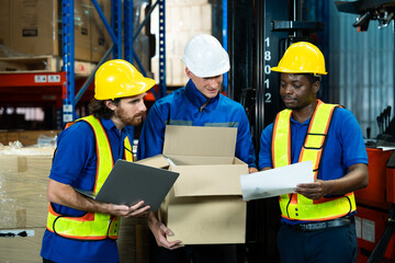 Group of three warehouse workers from different ethnic discussing warehouse operation holding cardboard boxes documents and laptop wearing safety helmets and reflective vests teamwork spirit