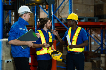 Three male warehouse workers from different ethnic backgrounds holding clipboard tablet and yellow safety helmet discussing teamwork strategy inside industrial warehouse with stacked boxes