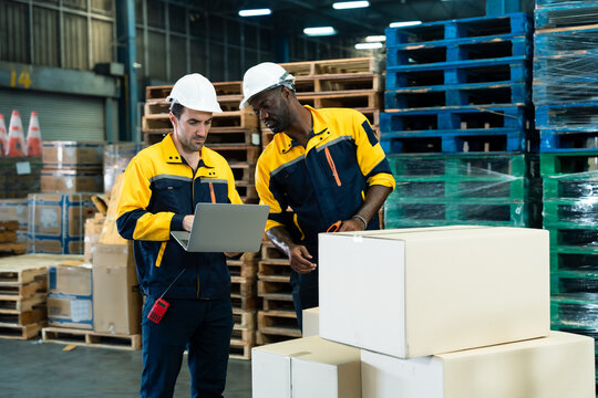 Caucasian and african male workers wearing hard hats using laptop and scanner to verify inventory and shipping data in busy warehouse zone full of stacked cartons and wooden pallets