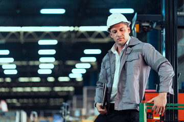Caucasian male technician in gray uniform wearing helmet leaning on forklift while holding laptop...