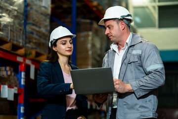 White adult female logistics staff using laptop beside storage racks during warehouse task operation, wearing safety helmet and navy blazer focused on screen, inside a logistics warehouse.