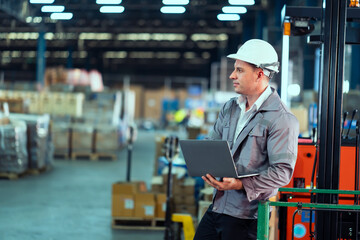 Caucasian male manager in gray uniform and white helmet holding laptop while analyzing production workflow inside spacious warehouse full of cargo and pallets, inside a logistics warehouse.