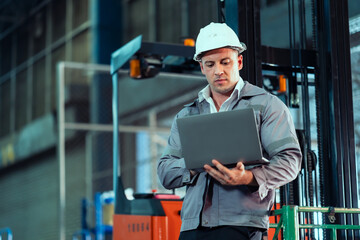 Caucasian male engineer wearing gray jacket and white helmet holding laptop inspecting forklift...