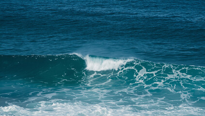 Fototapeta premium A small ocean wave crests with white foam and spray on the deep blue sea.