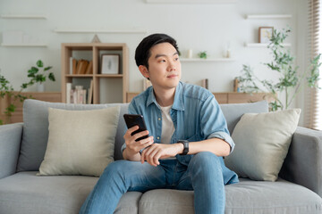 Obraz premium Smiling handsome young Asian man sitting on cozy couch, using funny mobile apps in living room. man at home, doing online shopping, messaging friends, posting on social media