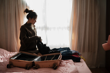 A woman sits on a bed, organizing clothes into an open suitcase, with sunlight filtering through...