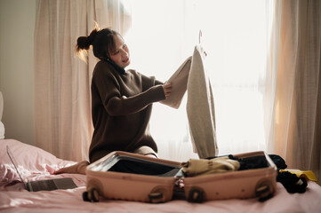 A woman is packing her suitcase on a bed while talking on the phone, surrounded by clothes and a...
