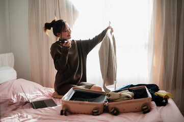 A young woman sits on a bed, smiling while holding a sweater, preparing her suitcase for travel...