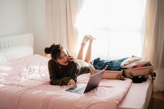 A young woman relaxes on a bed with a laptop, talking on the phone, surrounded by a suitcase and casual clothing in a cozy bedroom setting.