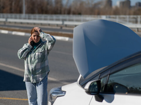 Caucasian woman cries and calls on the phone standing near the car with the hood open.  - Powered by Adobe
