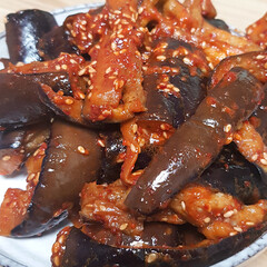 A close-up image of a dish featuring strips of eggplant mixed with a red savory sauce and sesame...