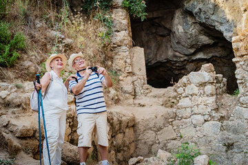 Senior couple tourists elderly people exploring ancient ruins with hiking poles, pensioners take pictures of the sights