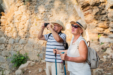 Senior couple tourists elderly people exploring ancient ruins with hiking poles, pensioners take pictures of the sights