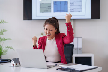 Asian adult woman celebrates real estate success using laptop at modern office with house plans...