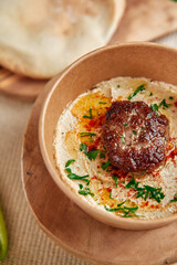 Satisfying bowl of creamy hummus topped with a perfectly grilled meat patty, garnished with fresh herbs, served alongside warm bread for a delicious and hearty meal.