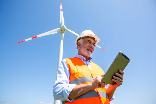 Experienced engineer inspects wind turbine operation with digital tablet under blue sky
