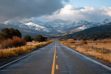 Naklejka premium Long straight road stretches toward snowy mountains under a dramatic sky with patches of sunlight illuminating the landscape in the late afternoon