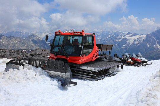 A Red Snowcat (Snow Groomer) And Snowmobiles Parked On A Glacier High In The Caucasus Mountains In Summer. Mountain Transport And Tourism.