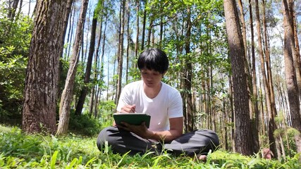 A young asian man wearing a white t-shirt and gray pants is drawing using a digital tablet while sitting on the grass in a lush pine forest in the morning. Remote work concept