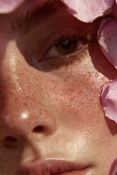 Close-up of a person's face adorned with pink flower petals, highlighting freckles and radiant skin in soft, natural light. Beauty in nature.