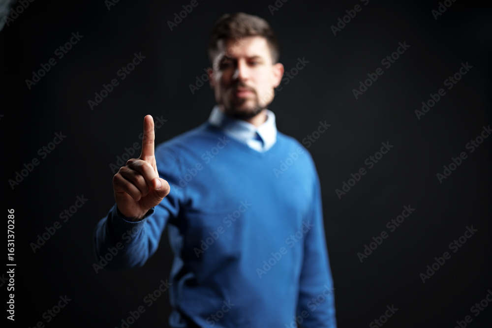 Poster man in blue sweater raises hand with index finger pointing up in dark setting to convey a message