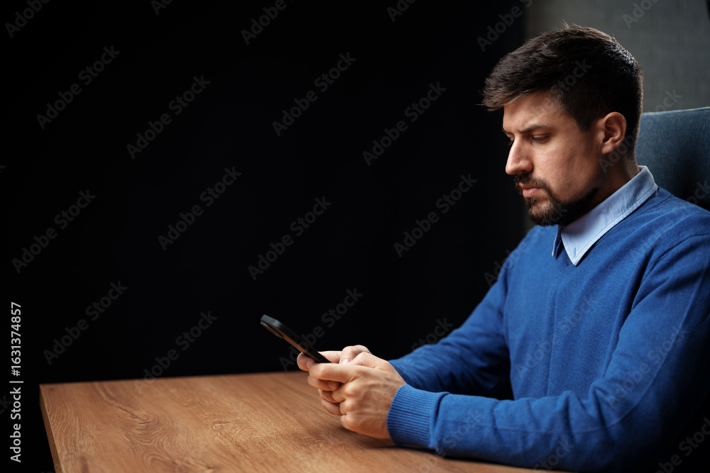 Poster man sitting at a desk using a smartphone in a dimly lit room with a contemplative expression