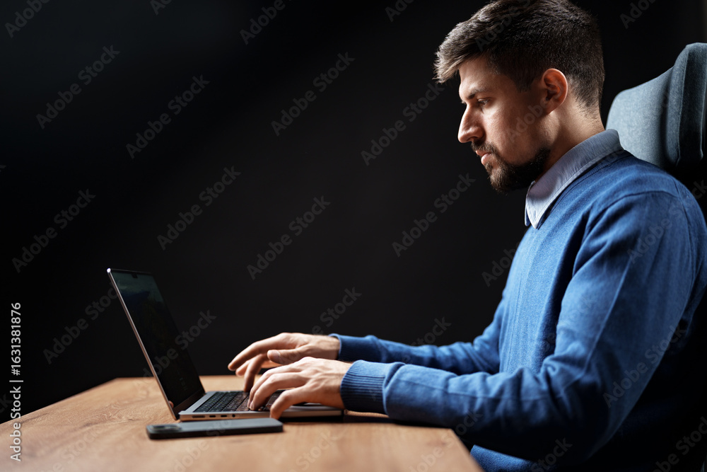 Poster man working intently on laptop at desk in a dimly lit environment during a focused session