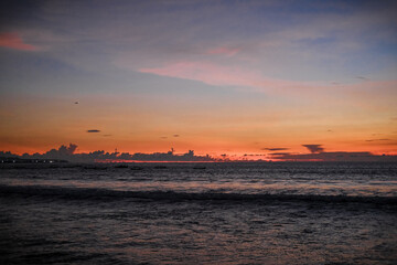 Obraz premium Golden sunset paints the sky over Kuta Beach, Bali, blending warm orange and purple hues above calm ocean waves, creating a serene and breathtaking view along the horizon of this tropical coast.