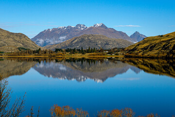 Obraz premium Snow-dusted mountains reflect perfectly in a calm lake under a clear blue sky near Queenstown, New Zealand, with trees and fields lining the peaceful shoreline.