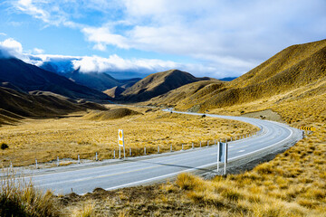 A winding road cuts through golden tussock hills under a moody sky in New Zealand’s South Island,...