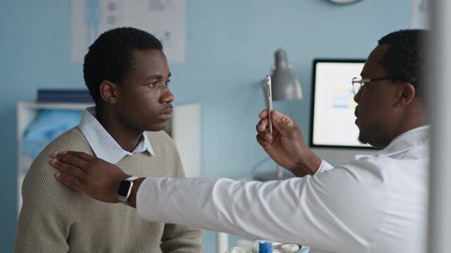 Side view waist up of Black male doctor performing eye movement and focus test by moving flashlight closer and farther from patient eyes, assessing reflexes and vision health during routine exam