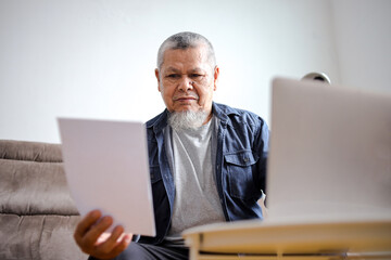 Senior Man With Casual Outfits Using Laptop And Noticed A Paper On His Hand